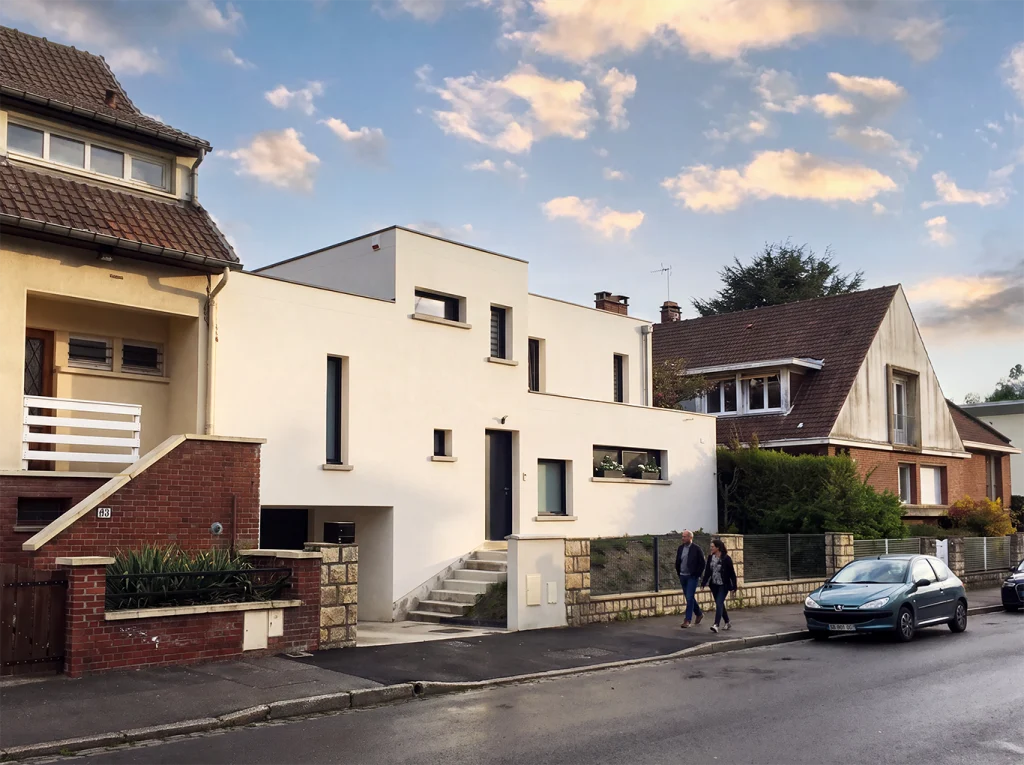 Vue oblique depuis la rue : escalier vers l’entrée surélevée et accès au garage en contrebas, entre les maisons voisines.
